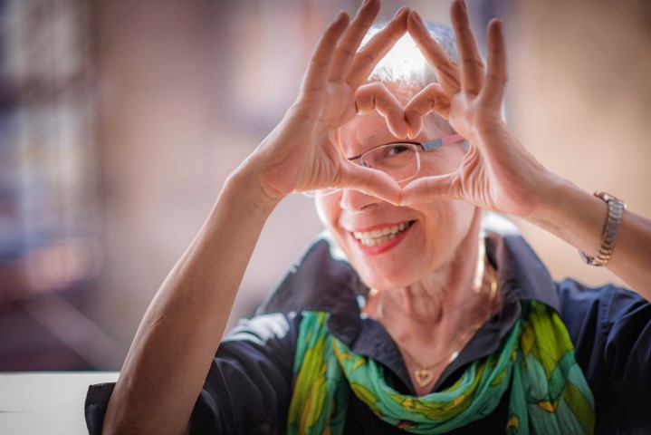 An older woman smiles and makes a heart symbol with her hands.