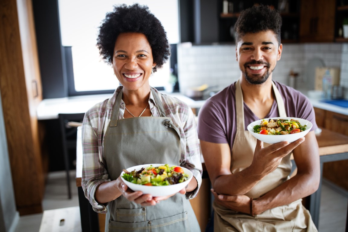 Couple holding bowls of healthy food in the kitchen and smiling.